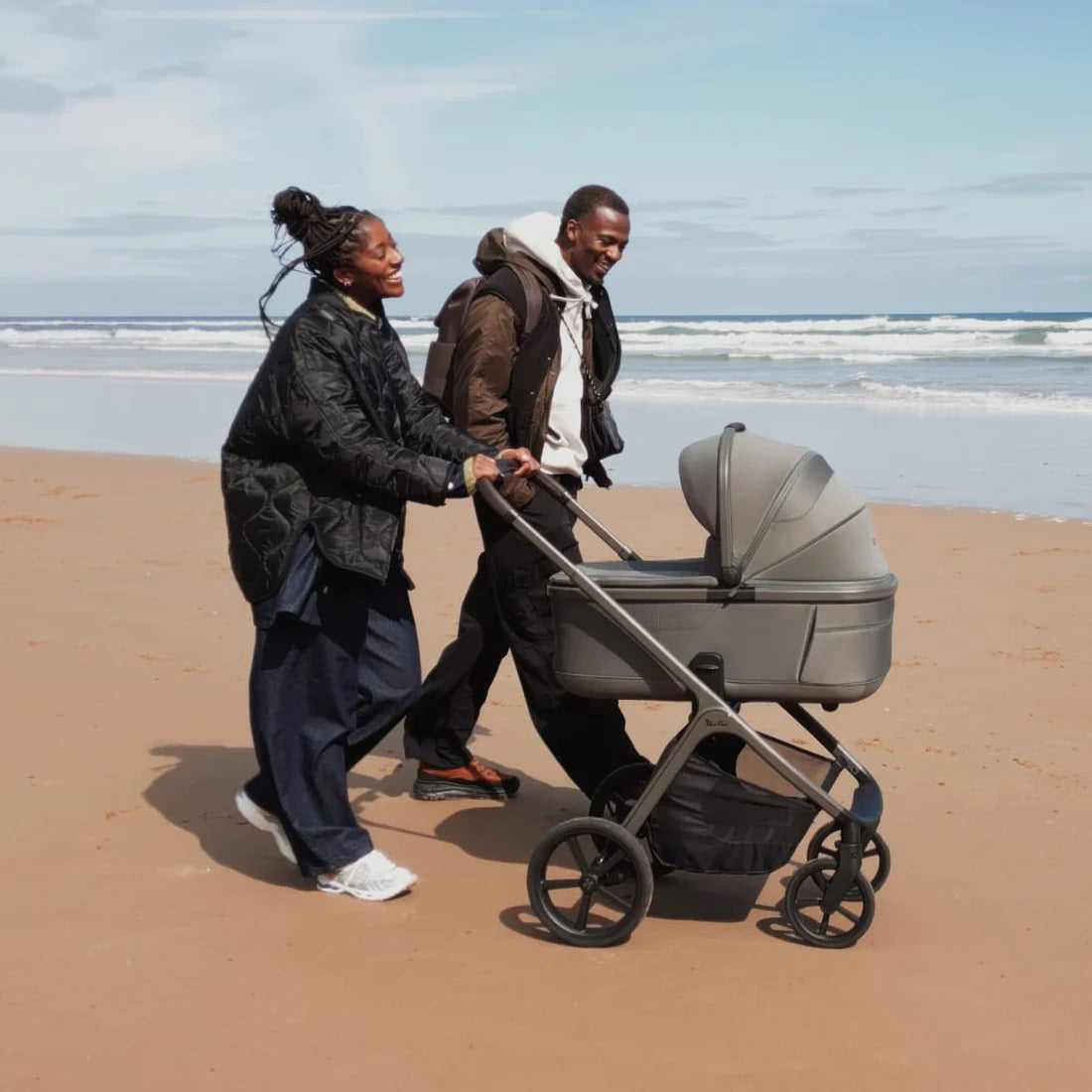 Two people pushing a stroller on a sandy beach with ocean waves in the background.